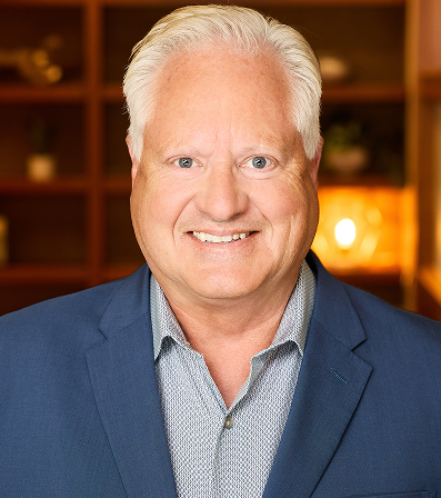 Smiling man in a blue blazer and patterned shirt against a blurred background.