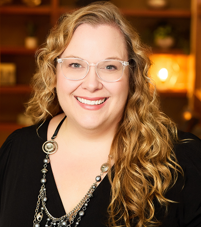 Woman with curly blonde hair and glasses, smiling, wearing a black top and silver necklace.