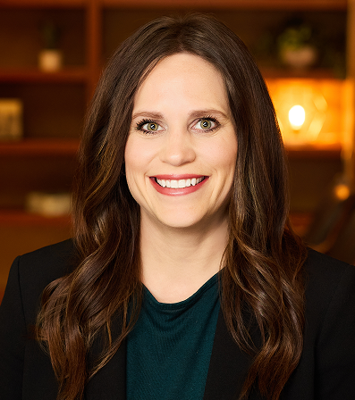 Woman with long brown hair smiles, wearing a black blazer and teal top in an office setting.