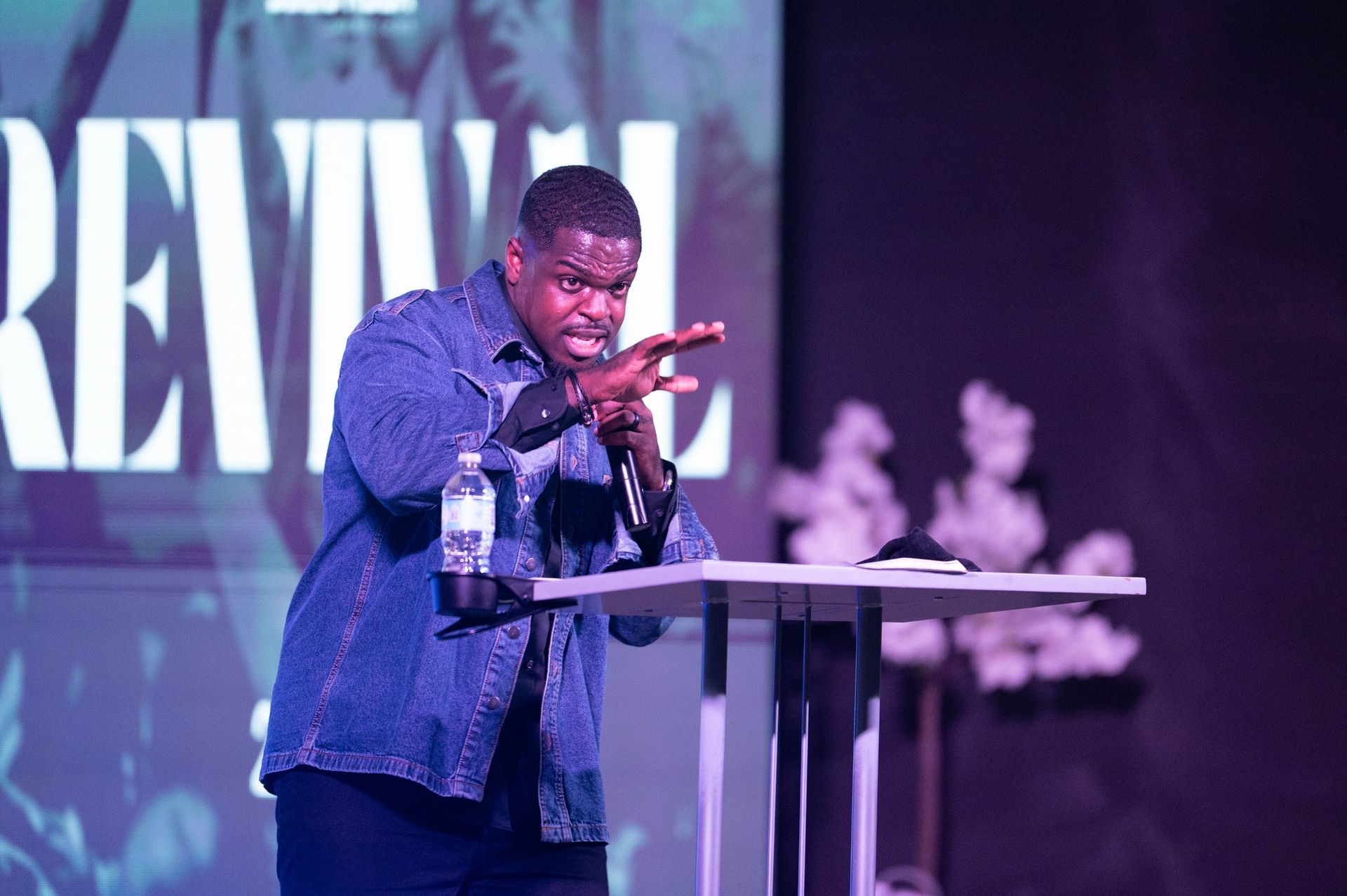 Man in denim jacket speaking at a podium, gesturing with hands, on stage