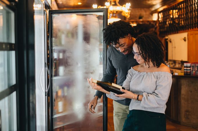 A man and a woman are looking at a bottle of wine in a refrigerator.
