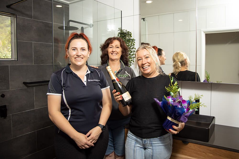 Three Women Are Standing in a Bathroom Holding a Bouquet of Flowers and a Bottle of Champagne — Omni Builders In Bungalow, QLD