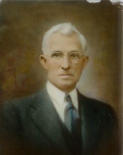 Portrait of a man in a dark suit, tie, and glasses, with white hair, set against a blurred background.