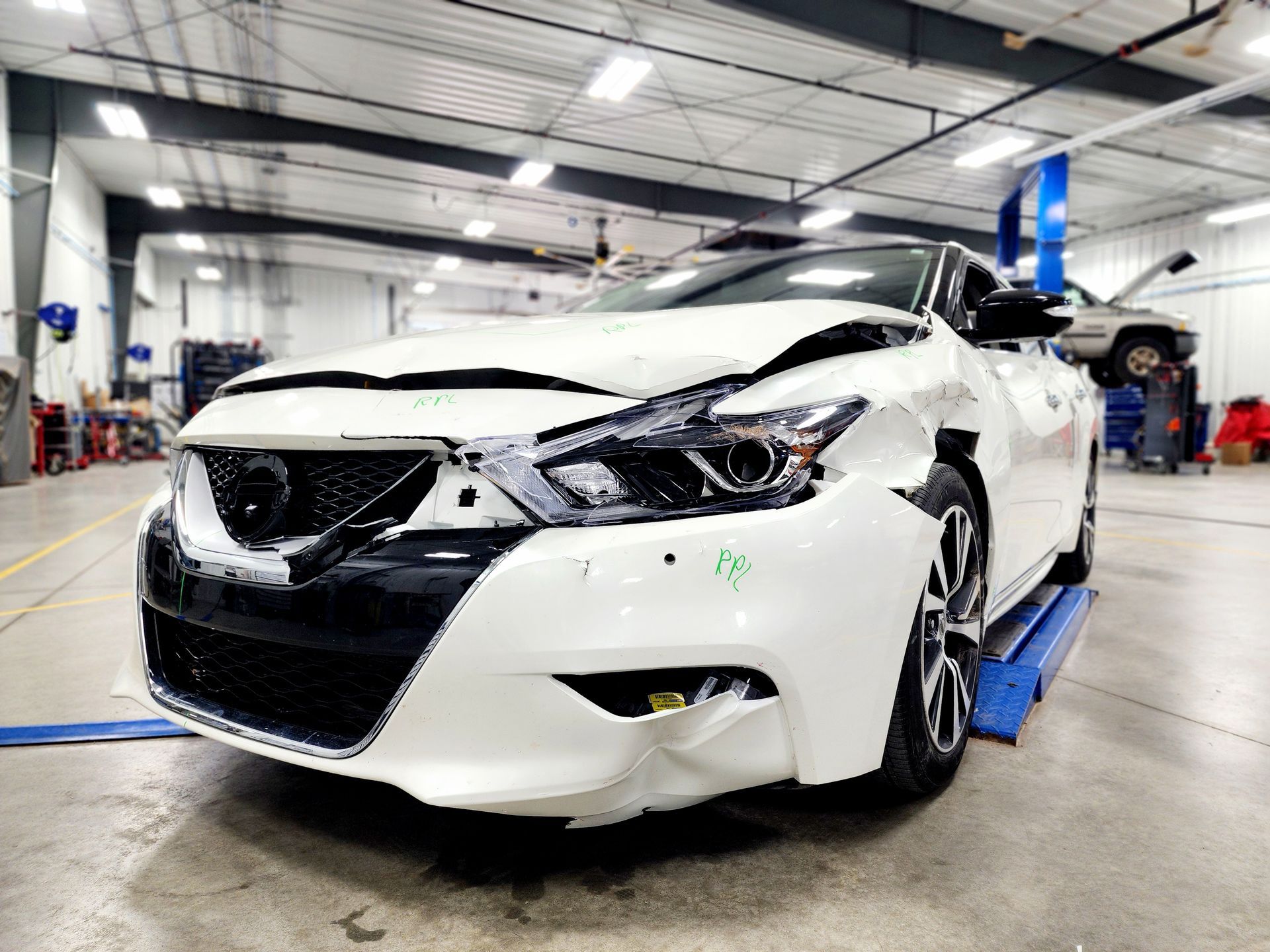 Damaged white sedan in an auto repair shop, front bumper and hood crumpled on a lift.