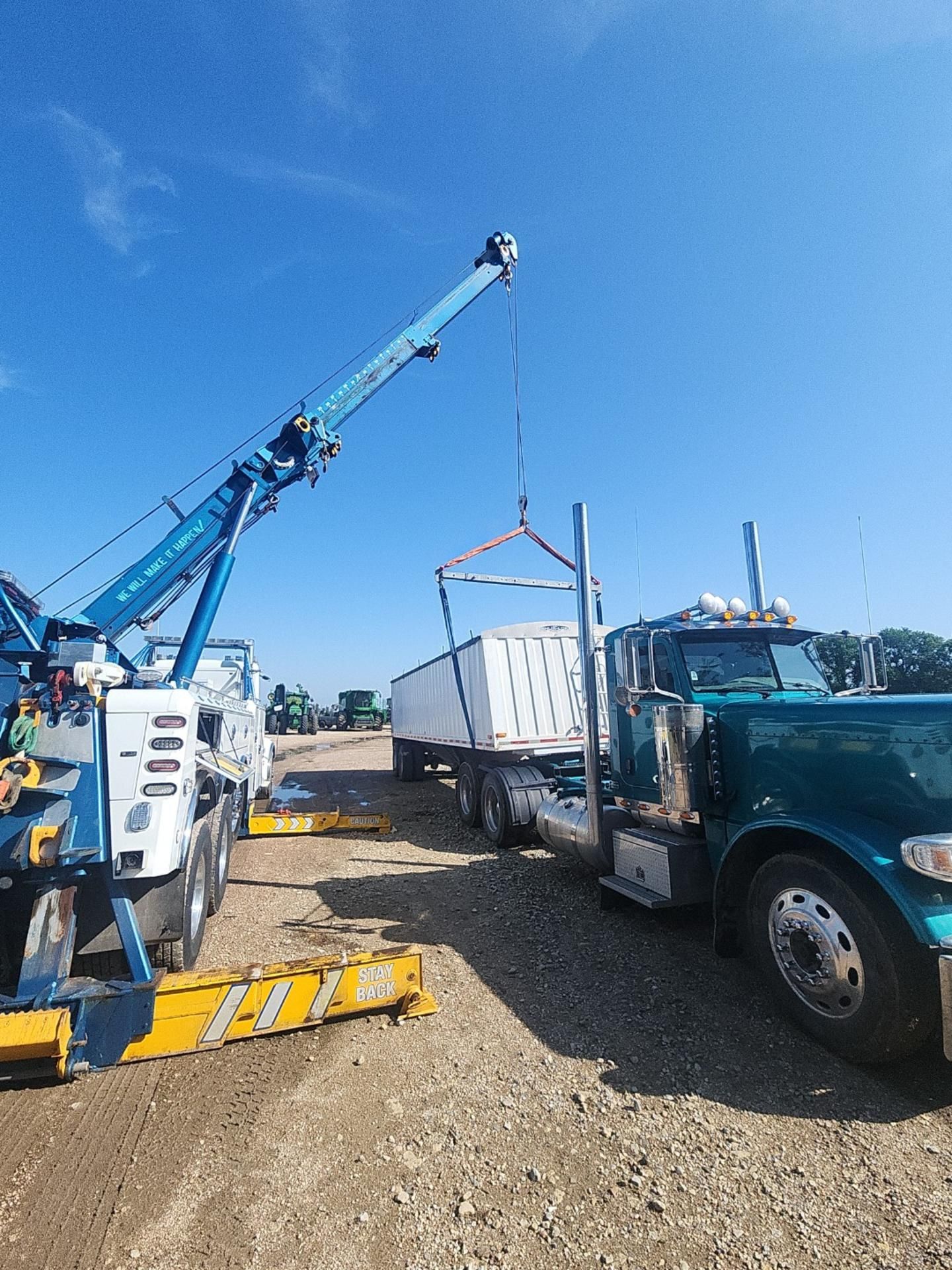 A blue tow truck lifting a white container from a teal semi truck on a gravel lot under a blue sky.
