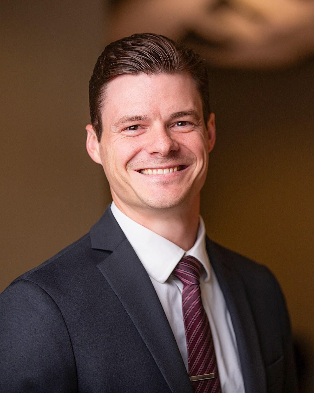 Smiling man in a dark suit and burgundy tie against a warm, blurred background
