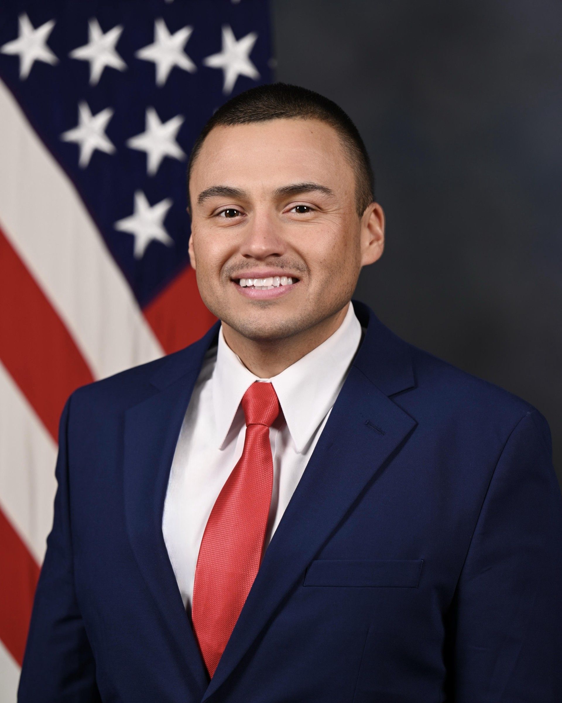 Portrait of a man in a navy suit and red tie, standing before an American flag backdrop.