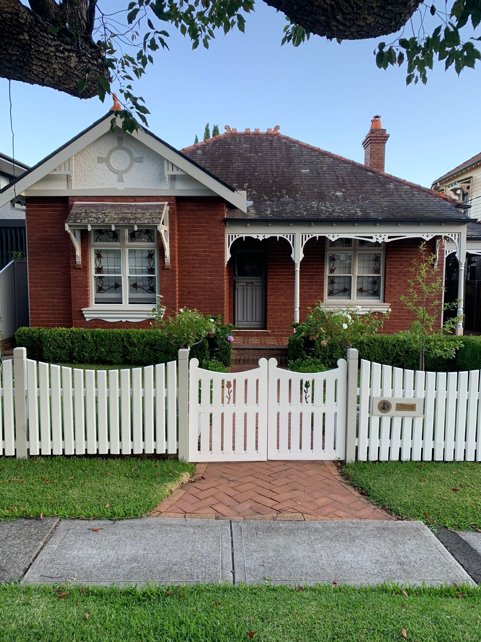 Red brick cottage with white picket fence, front porch, and trimmed hedges behind a sidewalk.