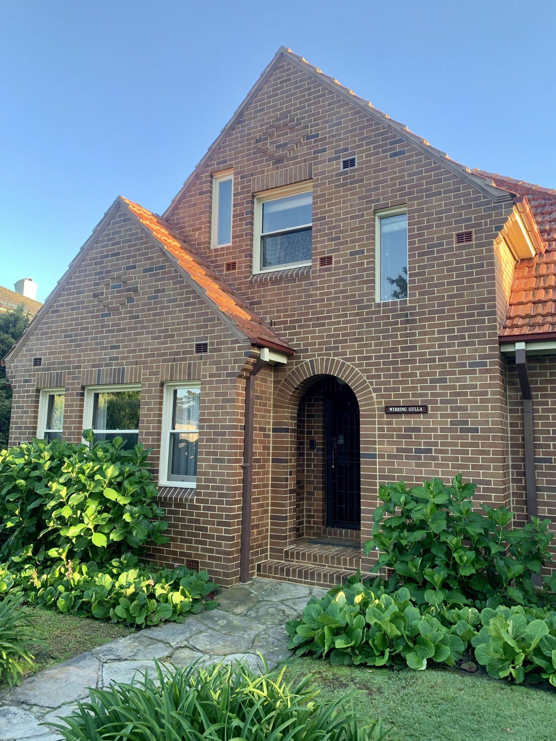 Brick house with steep gabled roof, arched doorway, and green shrubs in front