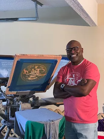 Man standing with arms crossed next to a screen printing machine, wearing a pink shirt, in a workshop.