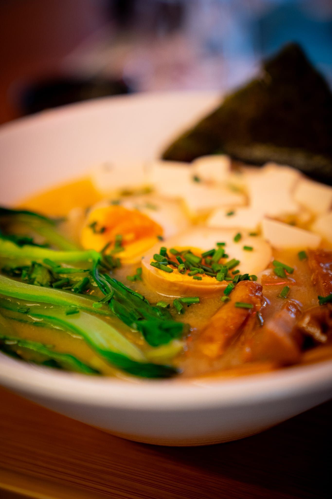Una ciotola di ramen con verdure verdi, uovo e alghe in un ristorante illuminato da luci soffuse.