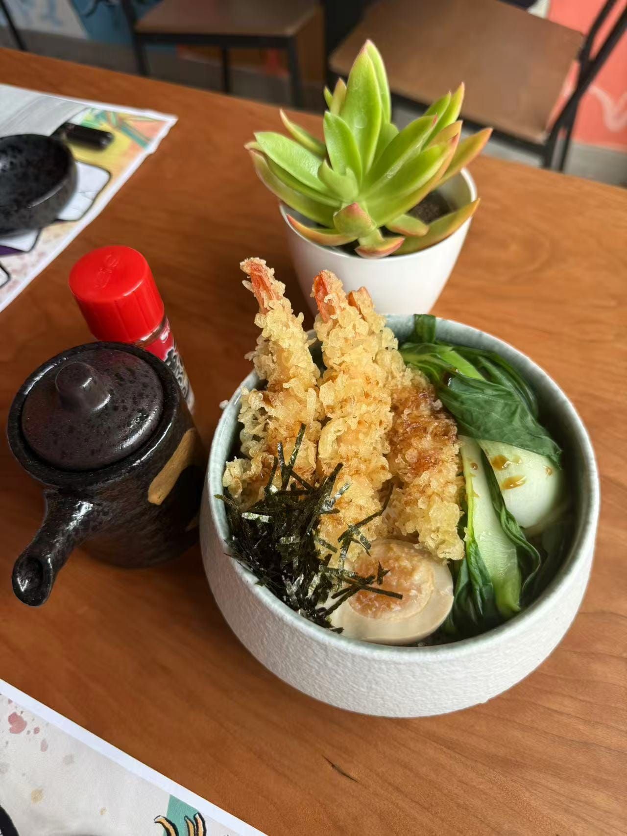 Ciotola di tempura giapponese con cetriolo, alghe, salsa di soia e una piccola pianta grassa in vaso su un tavolo di legno.