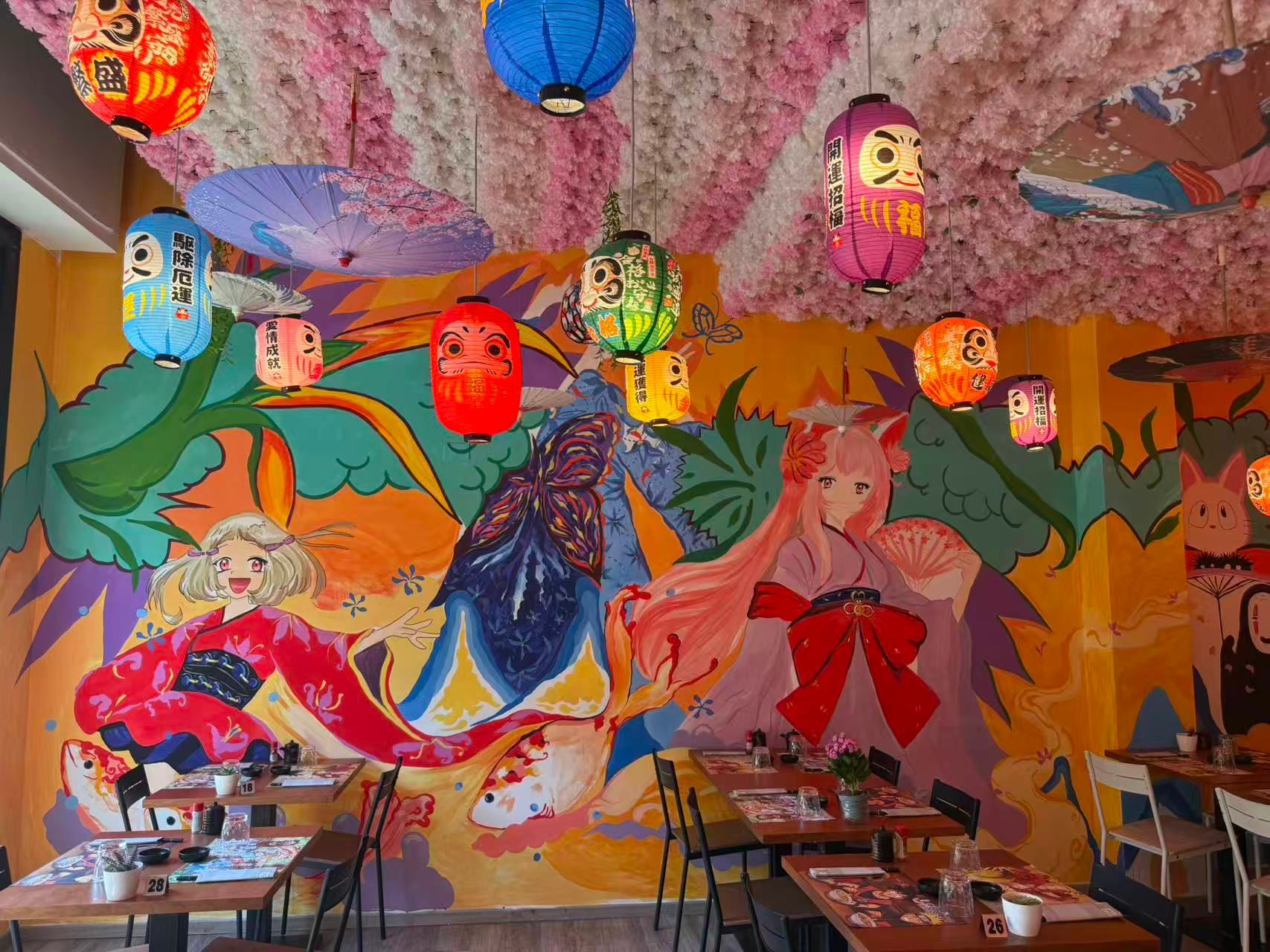 Caffè dai colori sgargianti, con lanterne sospese, decorazioni murali a forma di farfalla e tavoli sotto un soffitto con effetto materico.