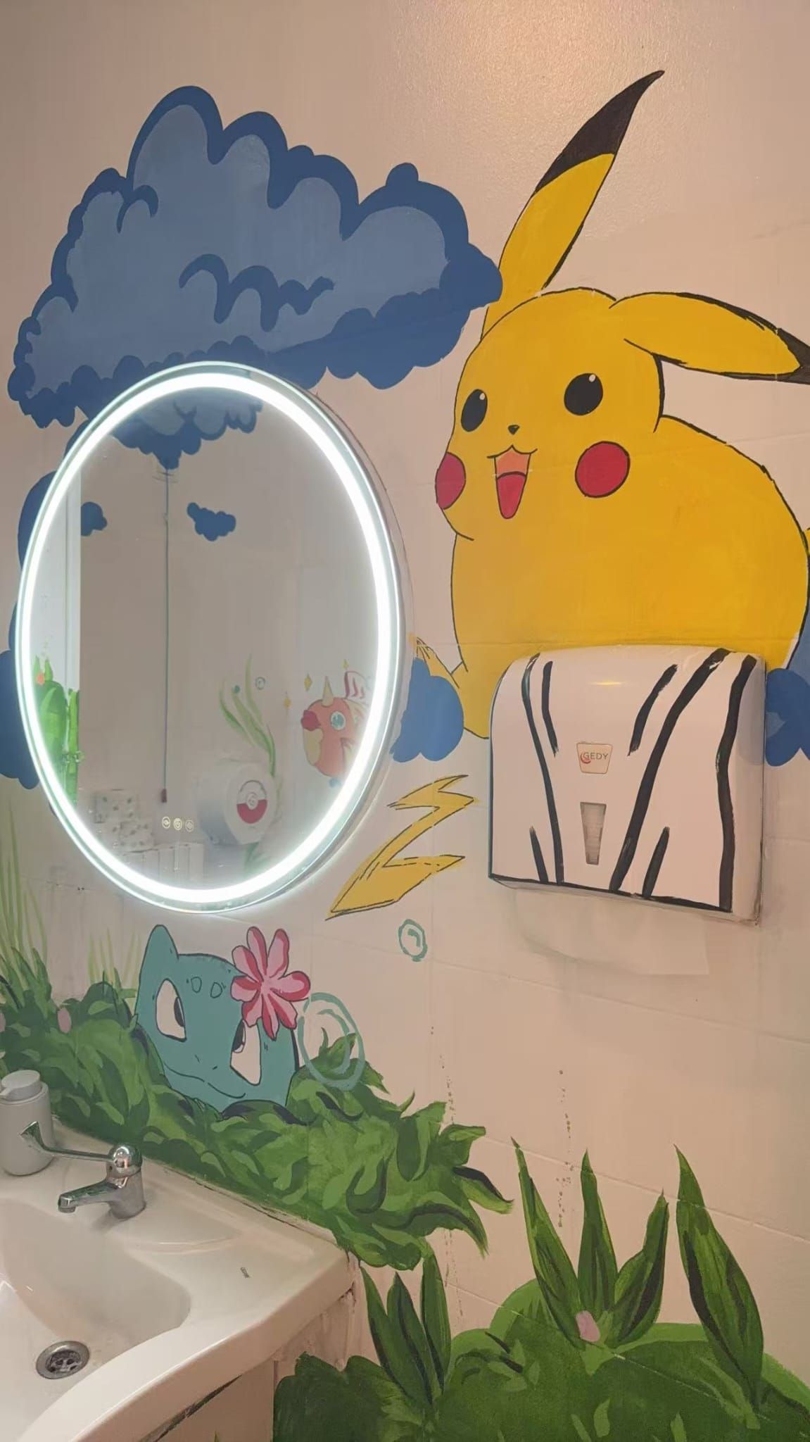 Un murale colorato con Pikachu accanto a uno specchio ovale sopra un lavandino e delle piante in un bagno.