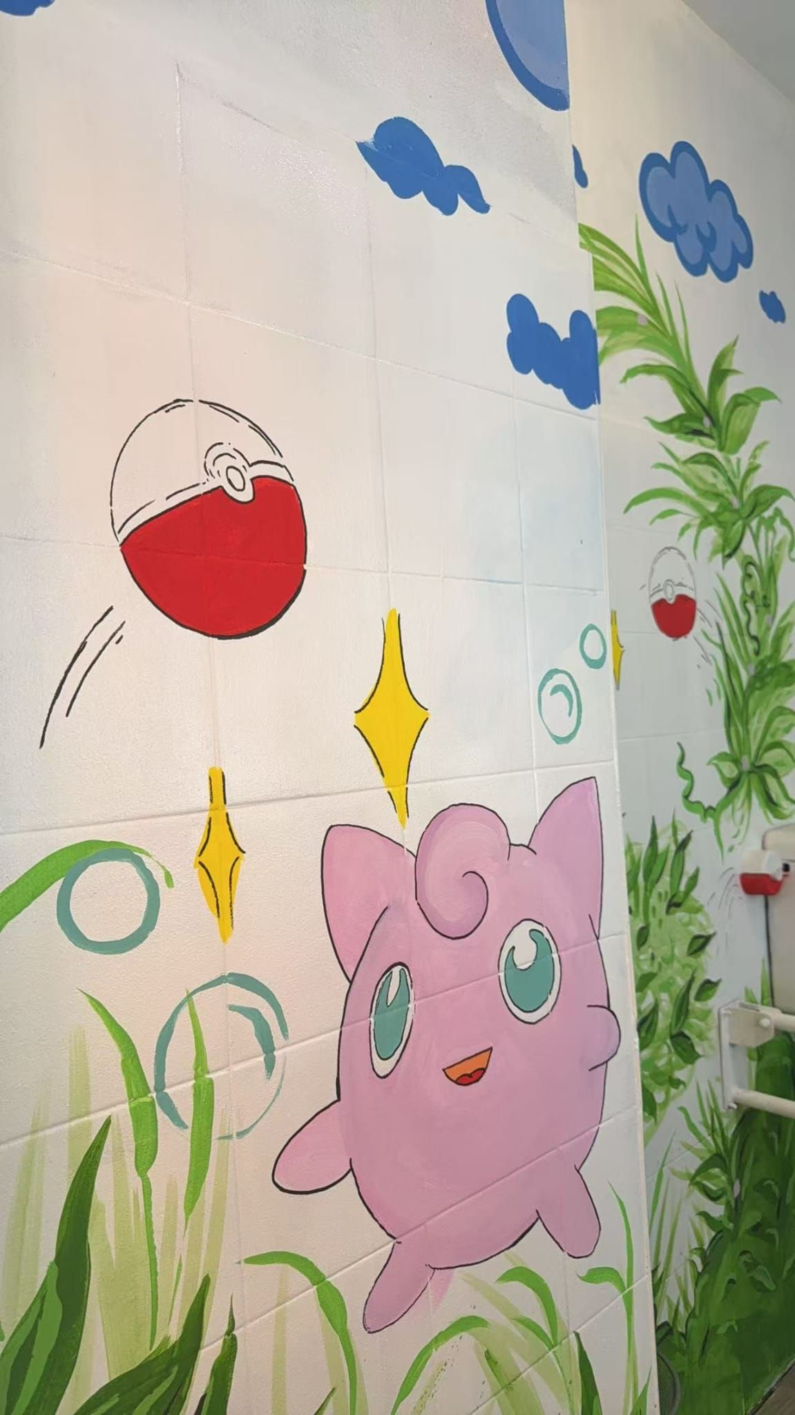Murale con un gatto rosa in stile cartone animato, piante verdi, nuvole blu e una palla rossa e bianca su uno sfondo bianco.