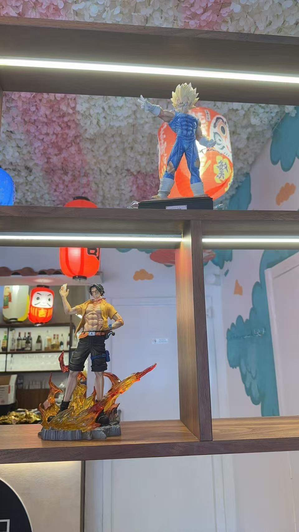 In una stanza, sono esposte delle miniature su scaffali di legno, tra cui una figura simile a un supereroe e una persona in piedi su una piattaforma.