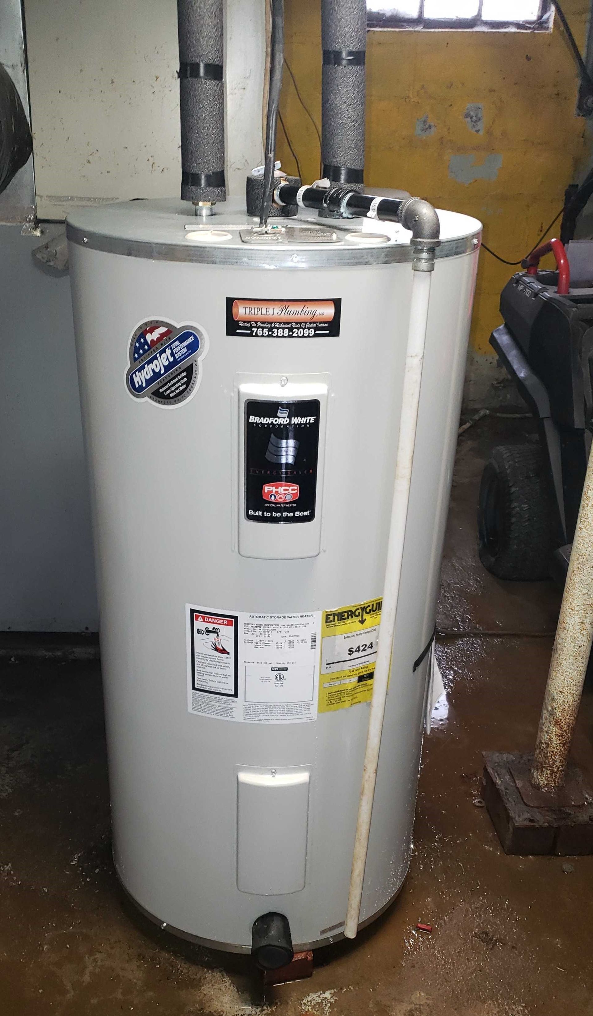 A white water heater is sitting in a basement next to a window.