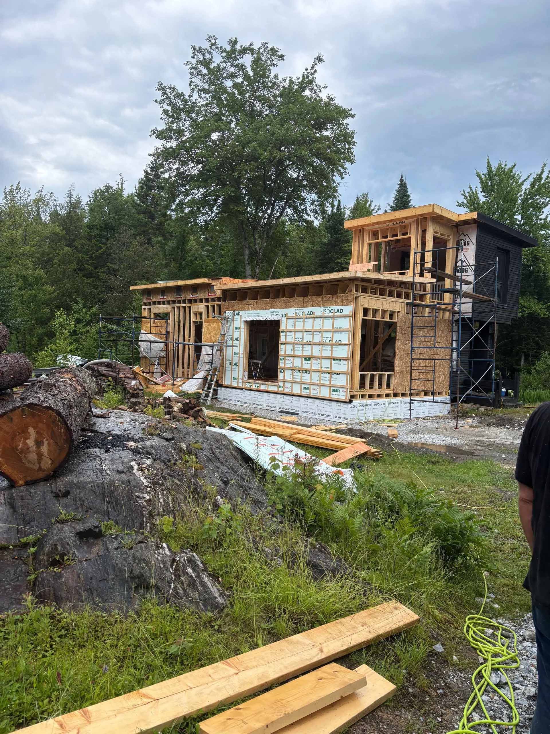 Une maison à ossature bois en construction dans une zone boisée, avec des poutres apparentes et un bardage partiellement installé.