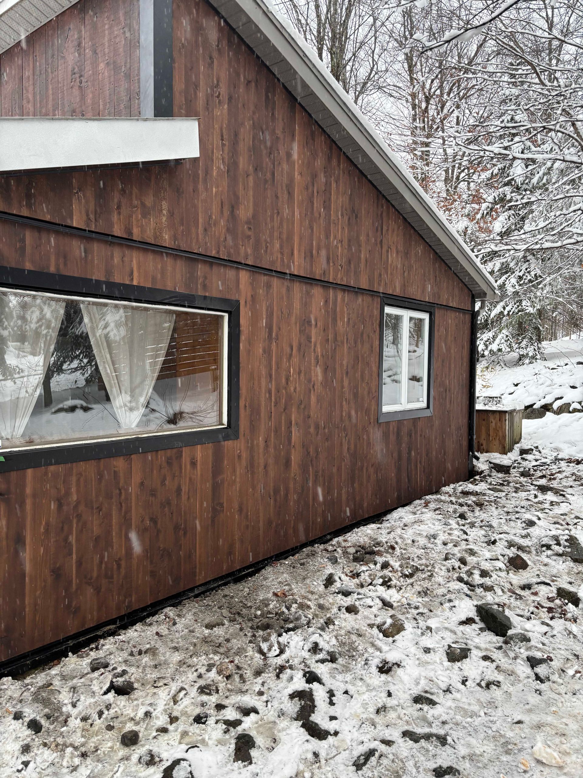 Vue latérale d'une cabane en bois brun avec deux fenêtres à cadre blanc, entourée de neige et d'arbres en hiver.