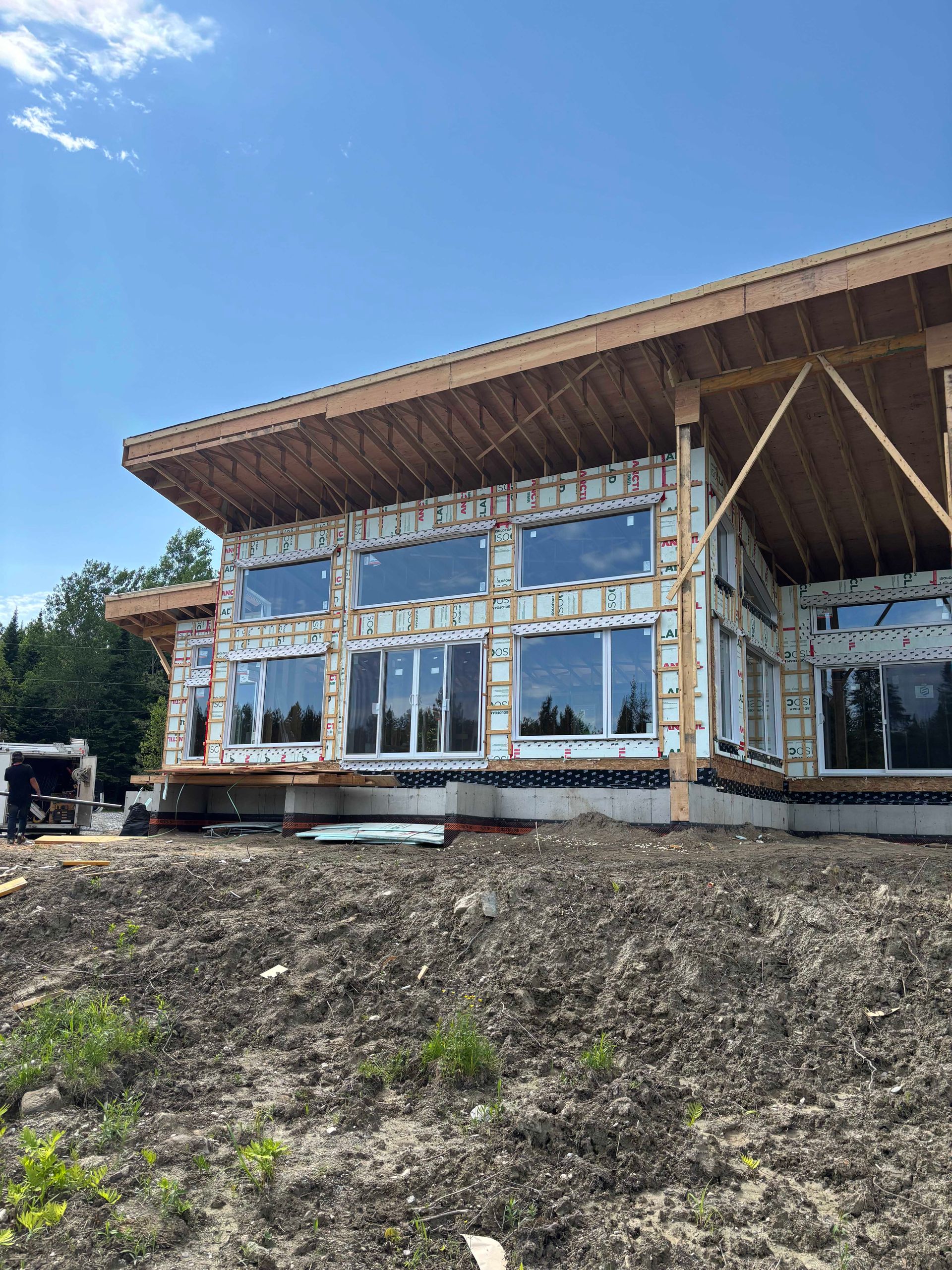 Une maison en construction, avec une charpente en bois apparente et de grandes fenêtres se détachant sur un ciel d'un bleu éclatant.