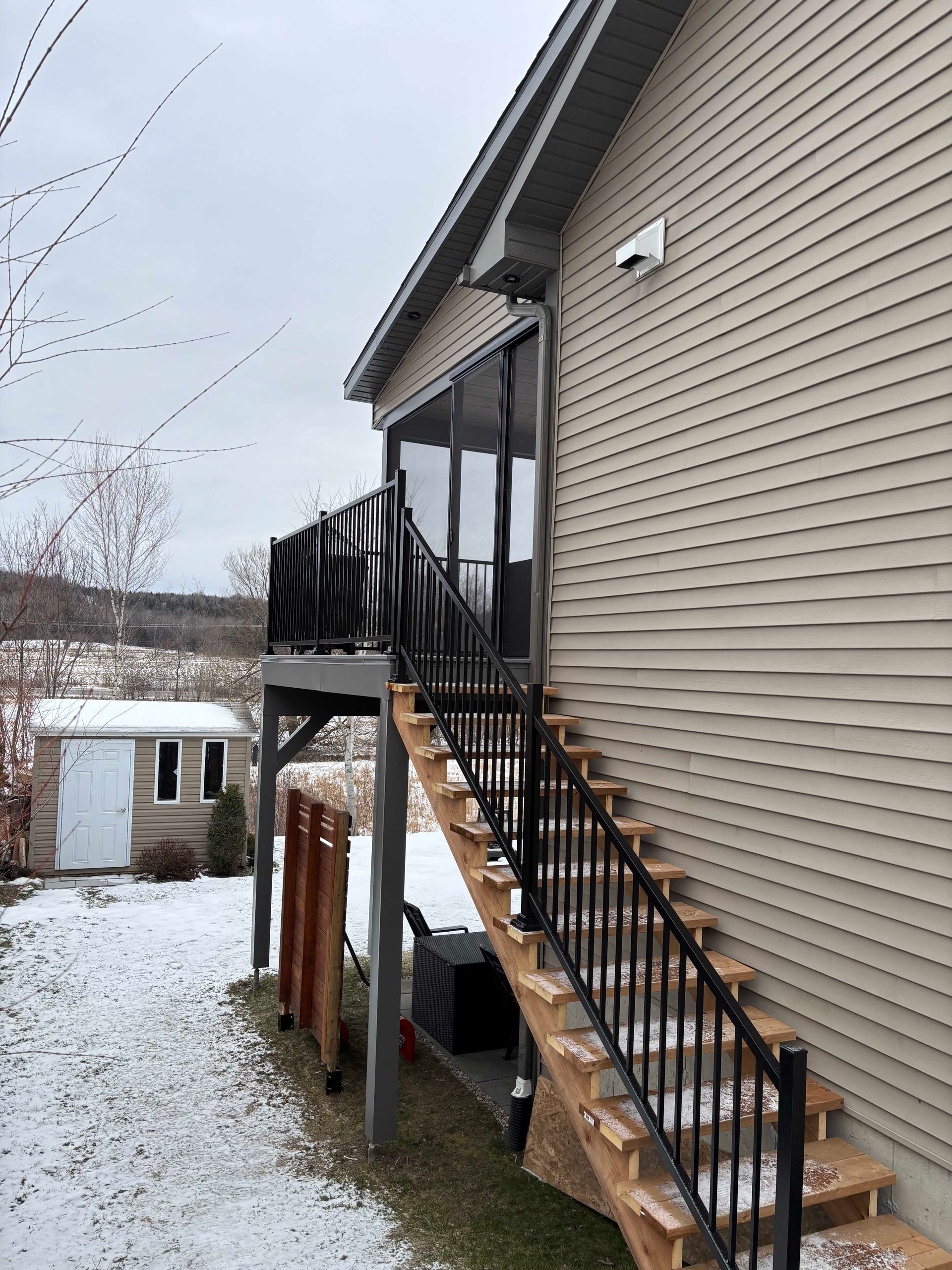 Vue extérieure d'une maison beige avec une terrasse en bois et un escalier en métal noir descendant vers une cour enneigée.