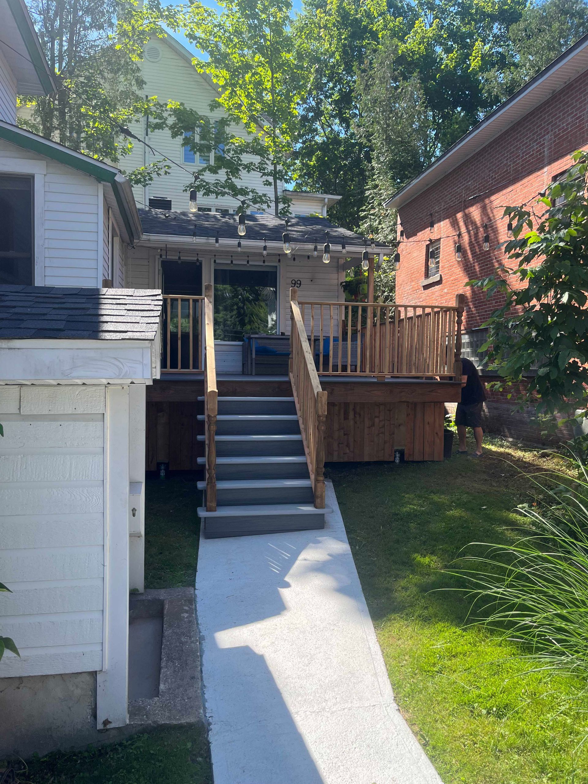Une terrasse en bois avec des escaliers mène à l'entrée arrière de la maison, reliée par une allée en béton blanc dans une cour verdoyante.