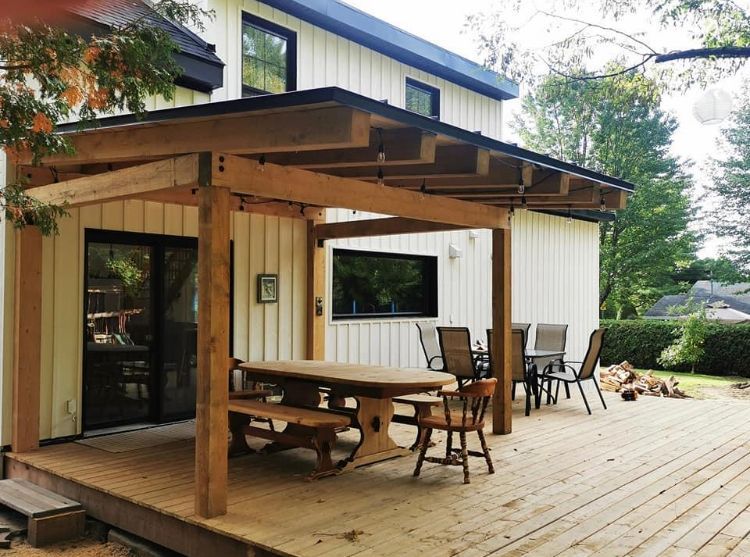 Une pergola en bois surplombe une terrasse où se trouvent une grande table à manger et des chaises, le tout adossé à une façade de maison aux tons clairs.