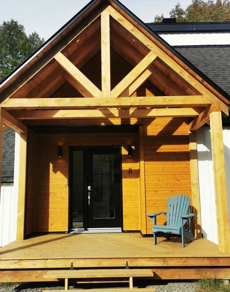 Une véranda à ossature bois avec un toit pointu, une porte d'entrée noire et une chaise Adirondack bleue sur une terrasse en bois.