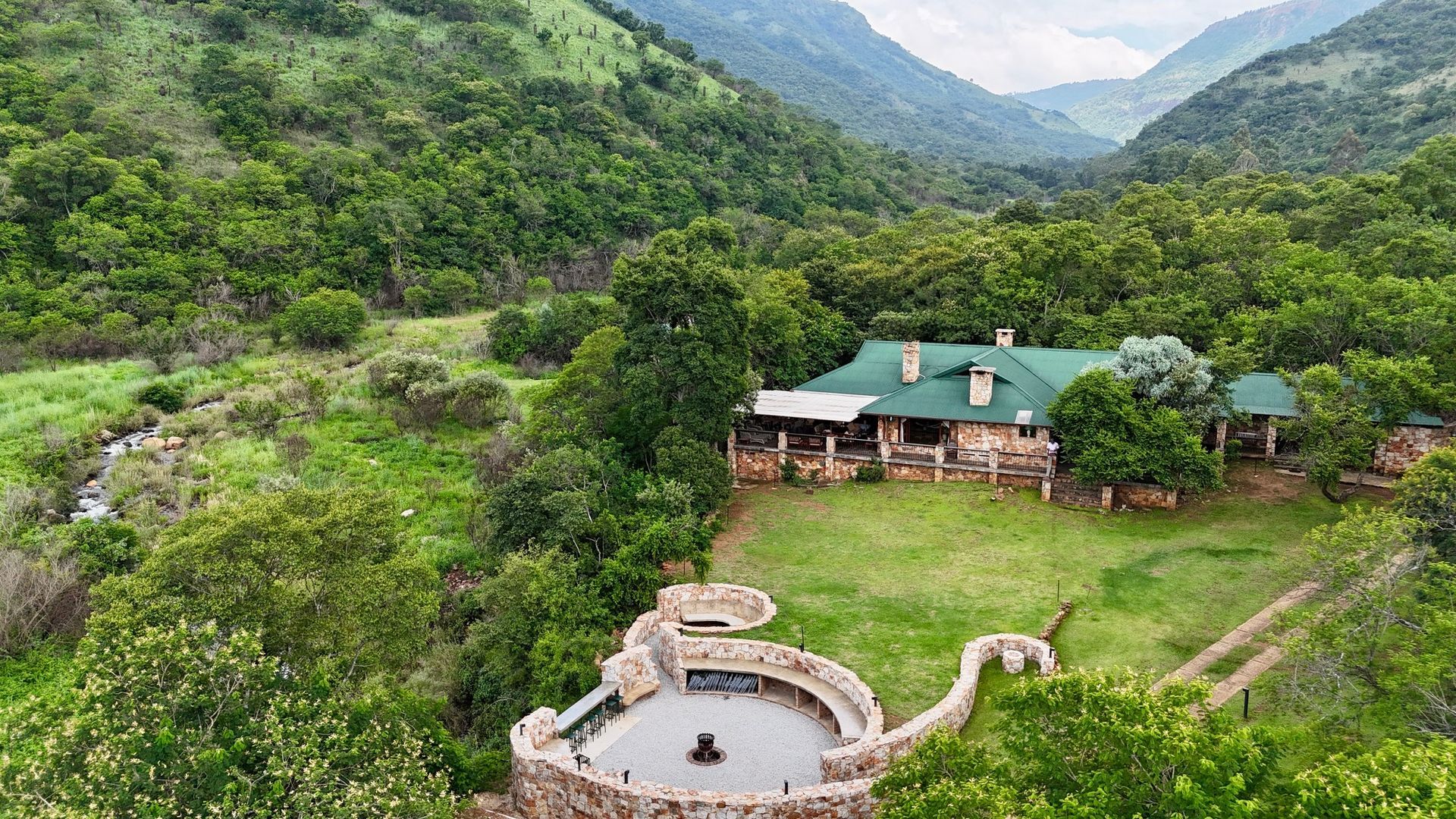 Lush mountain valley with a green-roof lodge and a circular stone boma nestled in dense greenery.