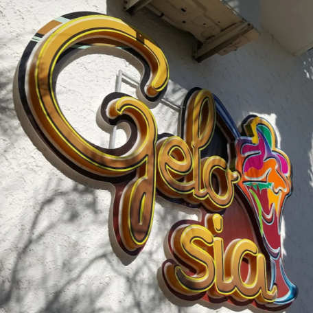 Insegna “Gelasia” color oro e rosso su una parete di stucco bianco, con una grafica a forma di volto colorato sulla destra.
