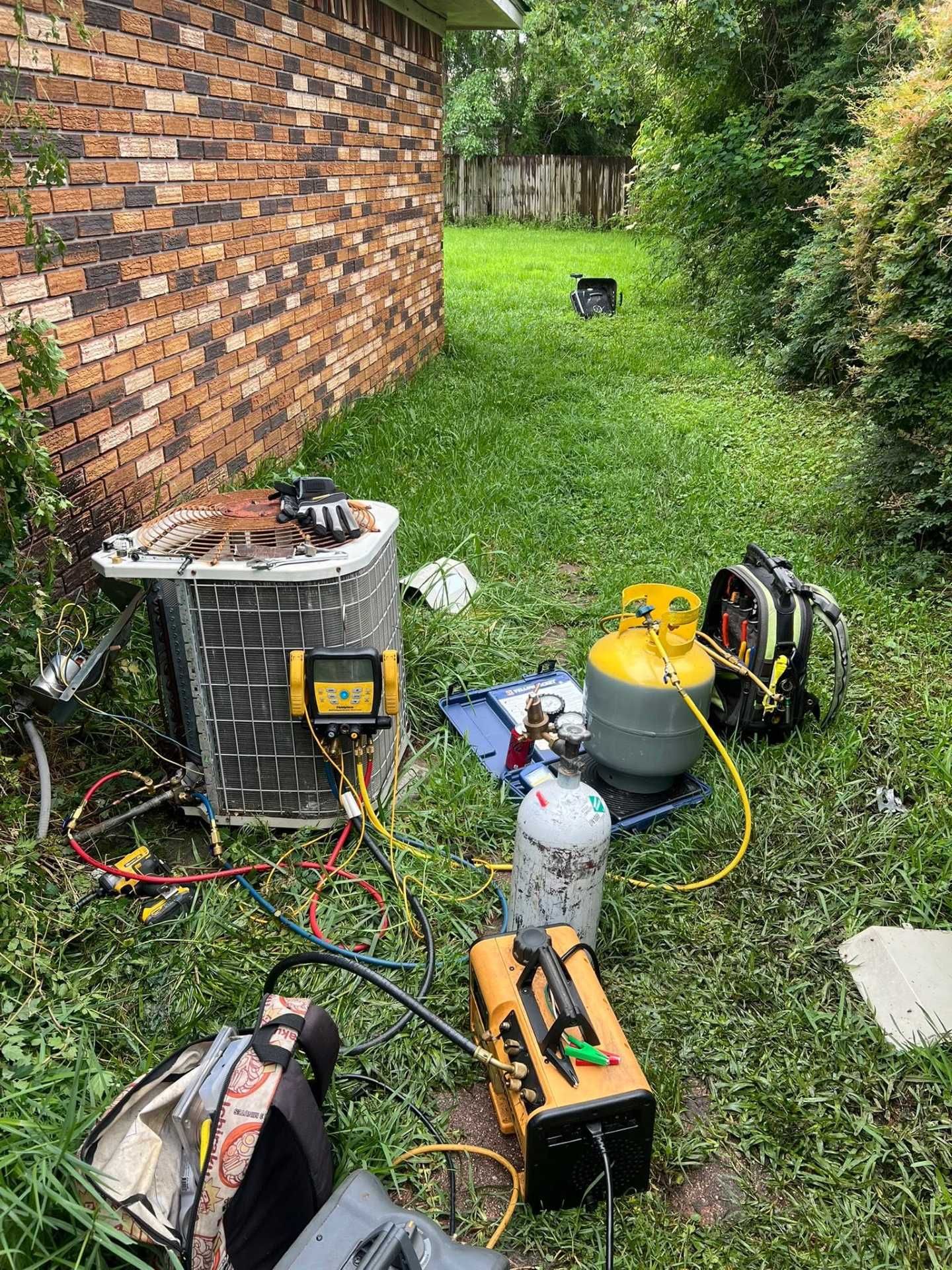 HVAC repair tools and equipment scattered on grass beside a brick house and outdoor AC unit.