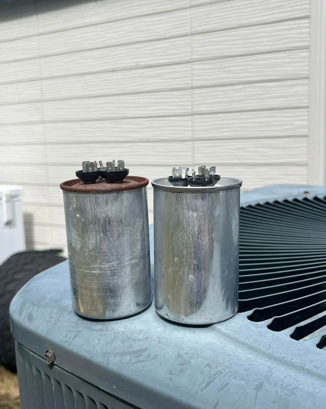 Two silver HVAC capacitors on top of an outdoor air conditioner unit, one rusted, one clean