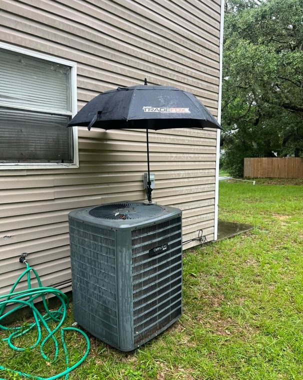 Black umbrella shading an outdoor air conditioner beside a beige house with a green hose on the grass