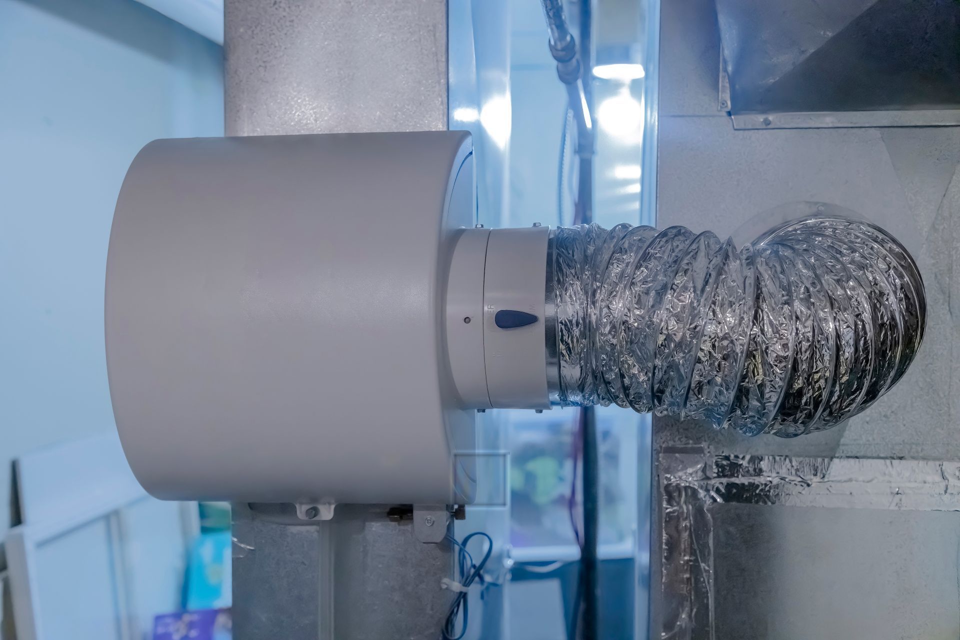 Wall-mounted exhaust fan with silver ducting in a room near a window