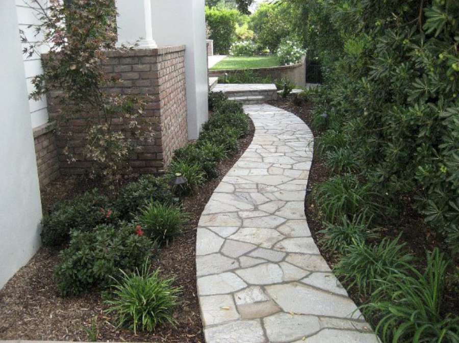 A stone walkway leading to a house surrounded by trees and bushes.