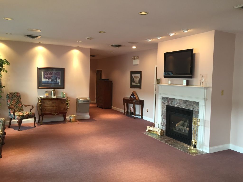 Spacious living room with a fireplace and TV, mauve carpet, and light pink walls. Furnishings include artwork, chairs, and decorative tables.