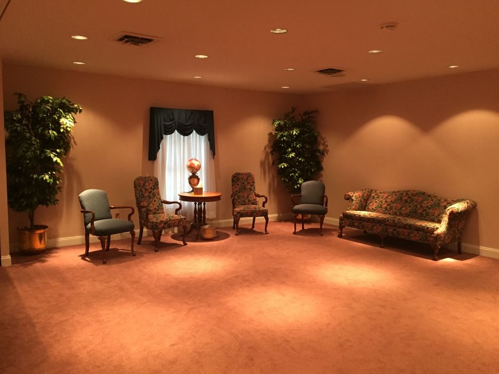 A softly lit room with tan walls and red carpet. It contains seating: a sofa, armchairs, and a round table near a window with a curtain. Potted plants are in corners.