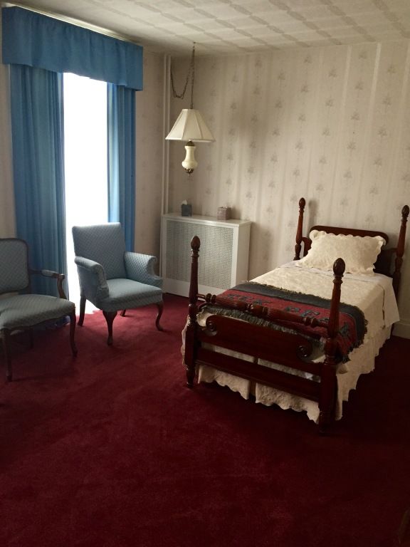 A bedroom with red carpet, a bed, two blue armchairs, and a window with blue curtains. Wallpaper is light colored with a star pattern.
