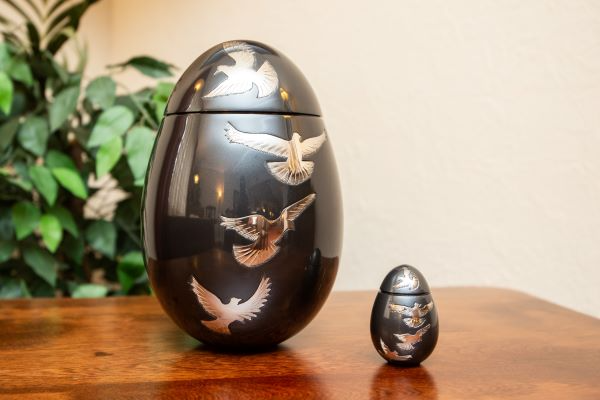Two egg-shaped black urns on a wooden table, decorated with silver doves. A small keepsake urn sits next to a larger one.