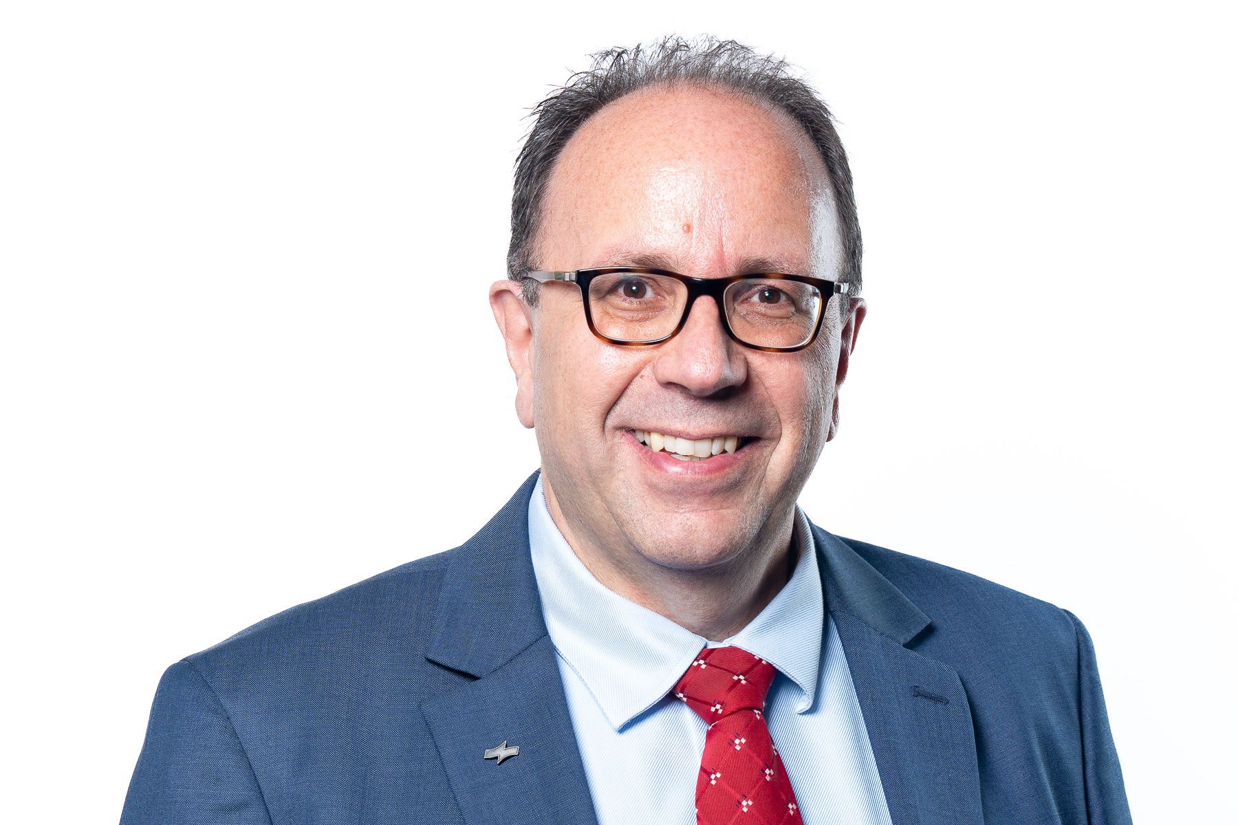 Studioaufnahme eines Mannes mit Brille, blauem Anzug und roter Krawatte, der vor einem weißen Hintergrund lächelt