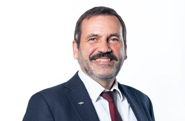 Porträt eines lächelnden Mannes in einem dunkelblauen Anzug, weißem Hemd und roter Krawatte vor weißem Hintergrund