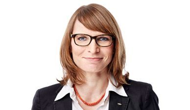 Porträt einer Frau mit Brille und roter Halskette, die einen schwarzen Blazer trägt, vor weißem Hintergrund