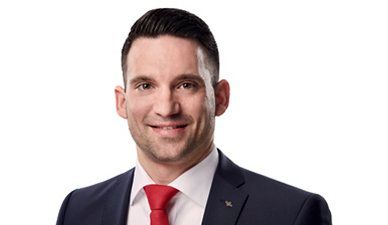 Porträt einer lächelnden Person in einem dunklen Anzug und roter Krawatte vor weißem Hintergrund