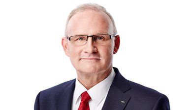 Porträt eines Mannes mit Brille, der einen dunkelblauen Anzug und eine rote Krawatte trägt, vor weißem Hintergrund
