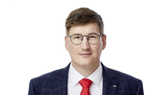 Porträt eines Mannes mit Brille, weißem Hemd, roter Krawatte und dunklem Blazer vor weißem Hintergrund