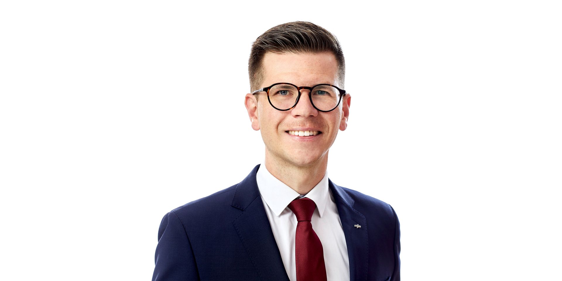 Porträt einer lächelnden Person in einem dunkelblauen Anzug, weißem Hemd und roter Krawatte vor weißem Hintergrund