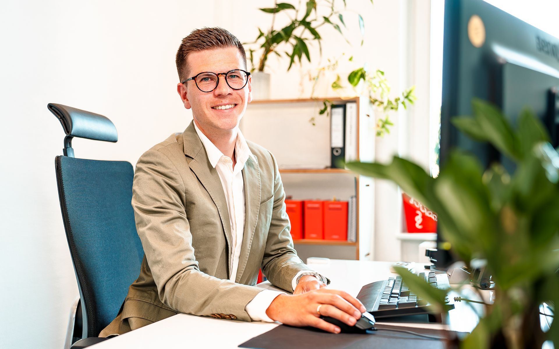 Ein lächelnder Geschäftsmann sitzt an einem Schreibtisch mit Computer in einem hellen Büro und tippt auf einer Tastatur.