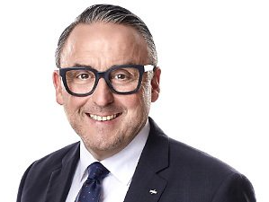 Lächelnder Mann in dunklem Anzug und Brille vor weißem Hintergrund