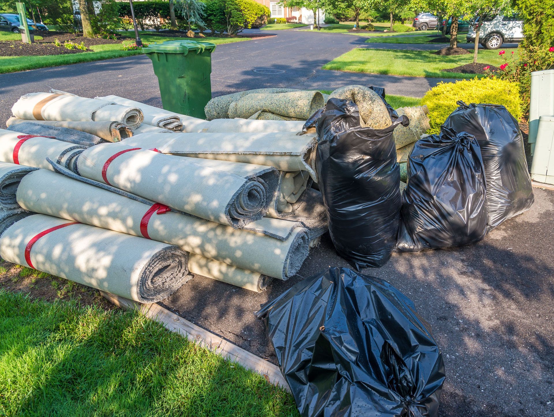 Rolled roofing materials and black trash bags piled on a driveway beside a suburban lawn