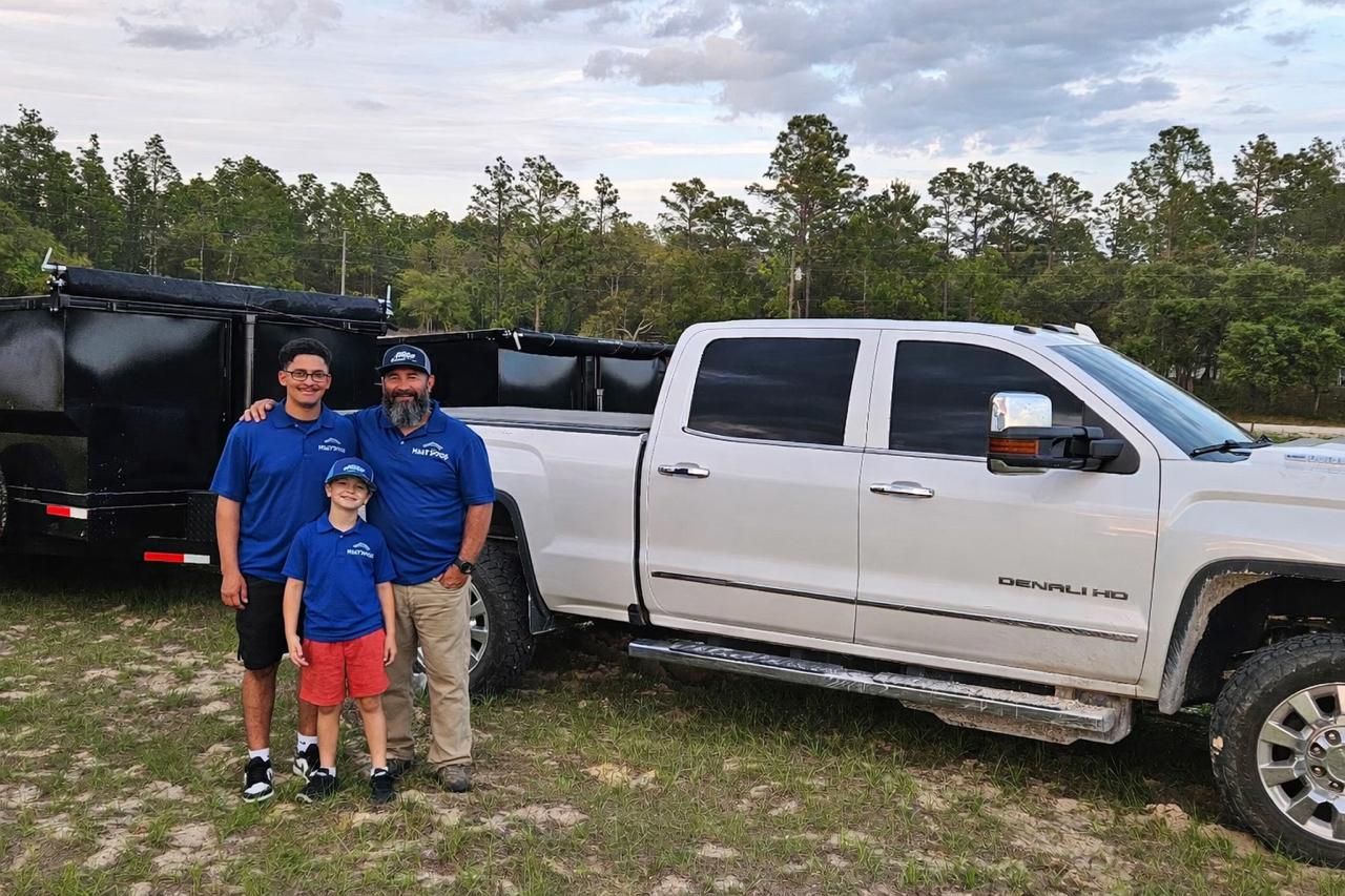 Three people standing beside a white pickup truck and black trailer in a grassy field